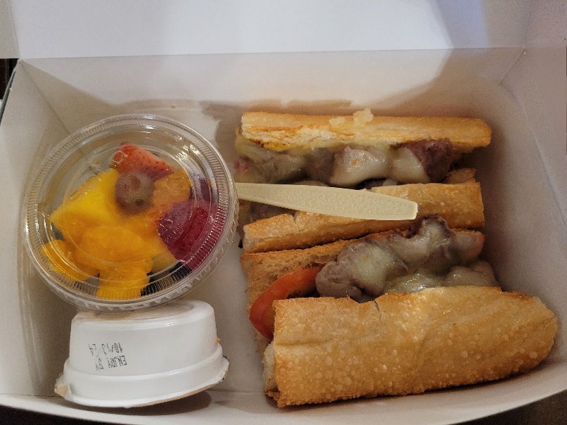 Beef sandwich, cup of fruit, and dressing container in a to go box.